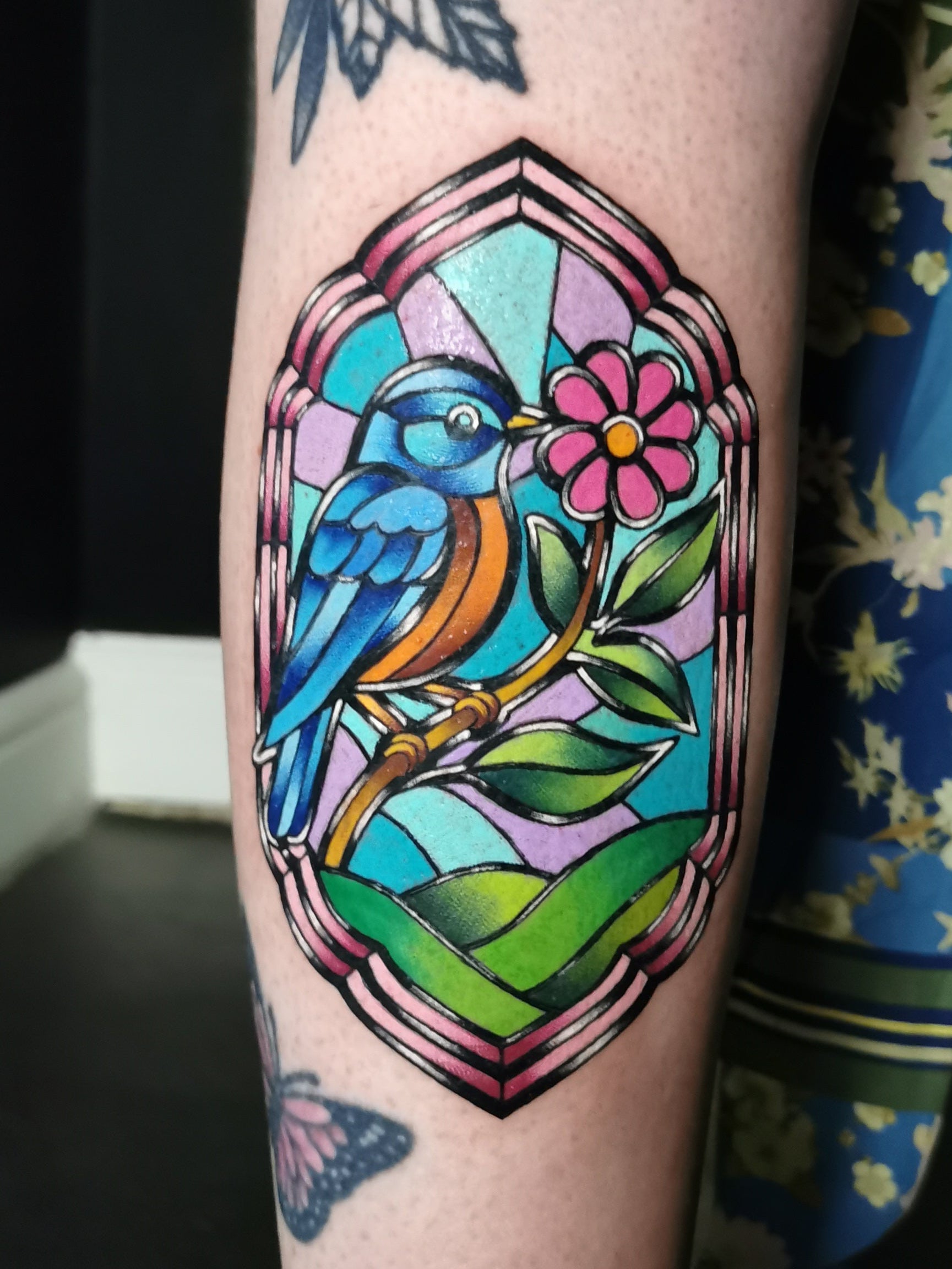Colorful tattoo of a bird and flower on a person's arm with a dark background. Stained glass window